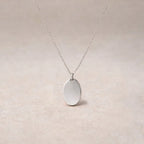 Collier "Tendresse", pendentif ovale personnalisable en acier inoxydable argenté, présenté sur un fond beige texturé.