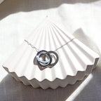 Collier "Je deviens" en acier argenté, avec trois cercles entrelacés personnalisables, posé sur un fond beige texturé.