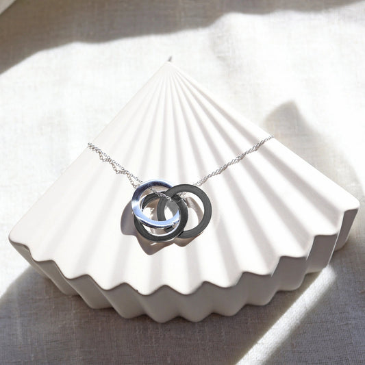 Collier "Je deviens" en acier argenté, avec trois cercles entrelacés personnalisables, posé sur un fond beige texturé.