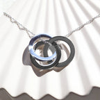 Zoom sur le collier "Je deviens" en acier argenté, avec trois cercles entrelacés personnalisables, posé sur un fond beige texturé.