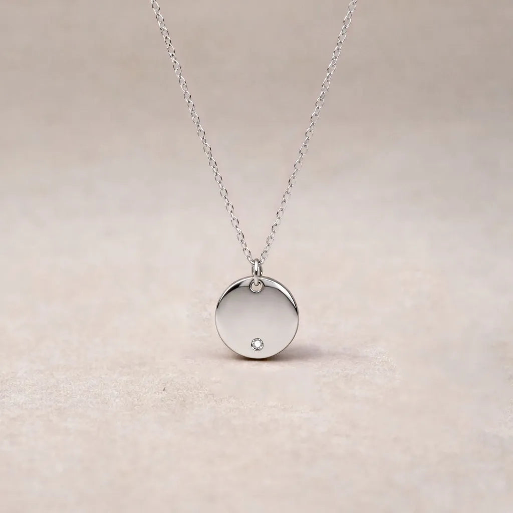 Zoom sur le collier personnalisable Halo avec médaille ronde brillante en acier argenté, posé sur un fond beige texturé.