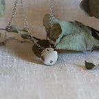 Collier personnalisable "Lumineuse", orné d'une médaille ronde brillante en acier argenté, posé sur un fond beige texturé.