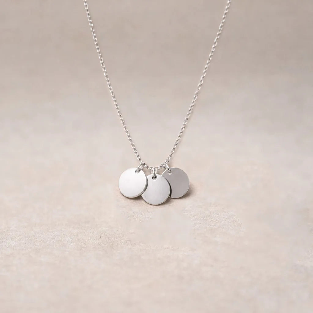 Collier "Inspiration" en acier inoxydable argenté, composé de trois médaillons personnalisables, présenté sur un fond beige texturé.