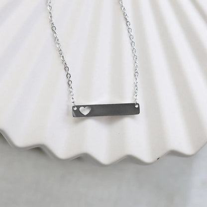 Collier plaque personnalisable en acier argenté avec cœur découpé, posé à plat sur un support texturé beige, photo en gros plan. 