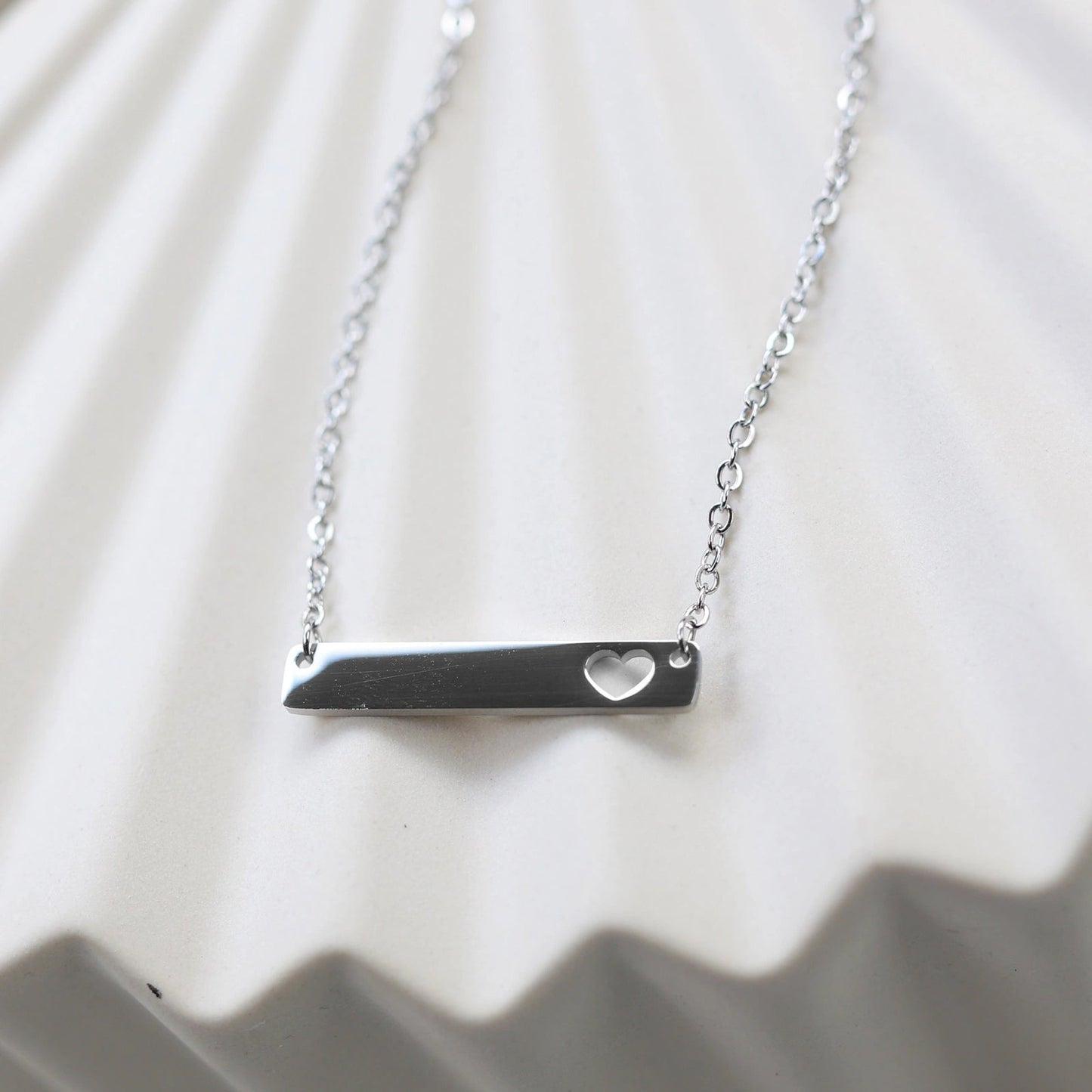 Collier plaque personnalisable en acier argenté avec cœur découpé, posé à plat sur un support texturé beige, photo en gros plan. 