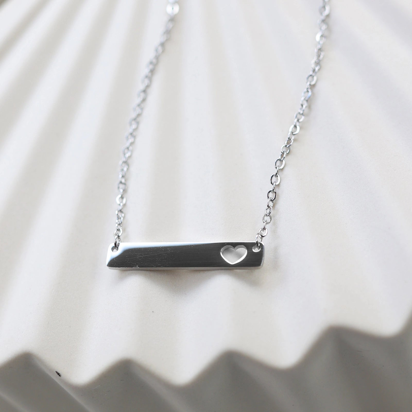 Collier plaque personnalisable en acier argenté avec cœur découpé, posé à plat sur un support texturé beige, photo en gros plan. 