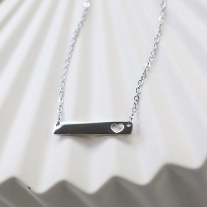 Collier plaque personnalisable en acier argenté avec cœur découpé, posé à plat sur un support texturé beige, photo en gros plan. 