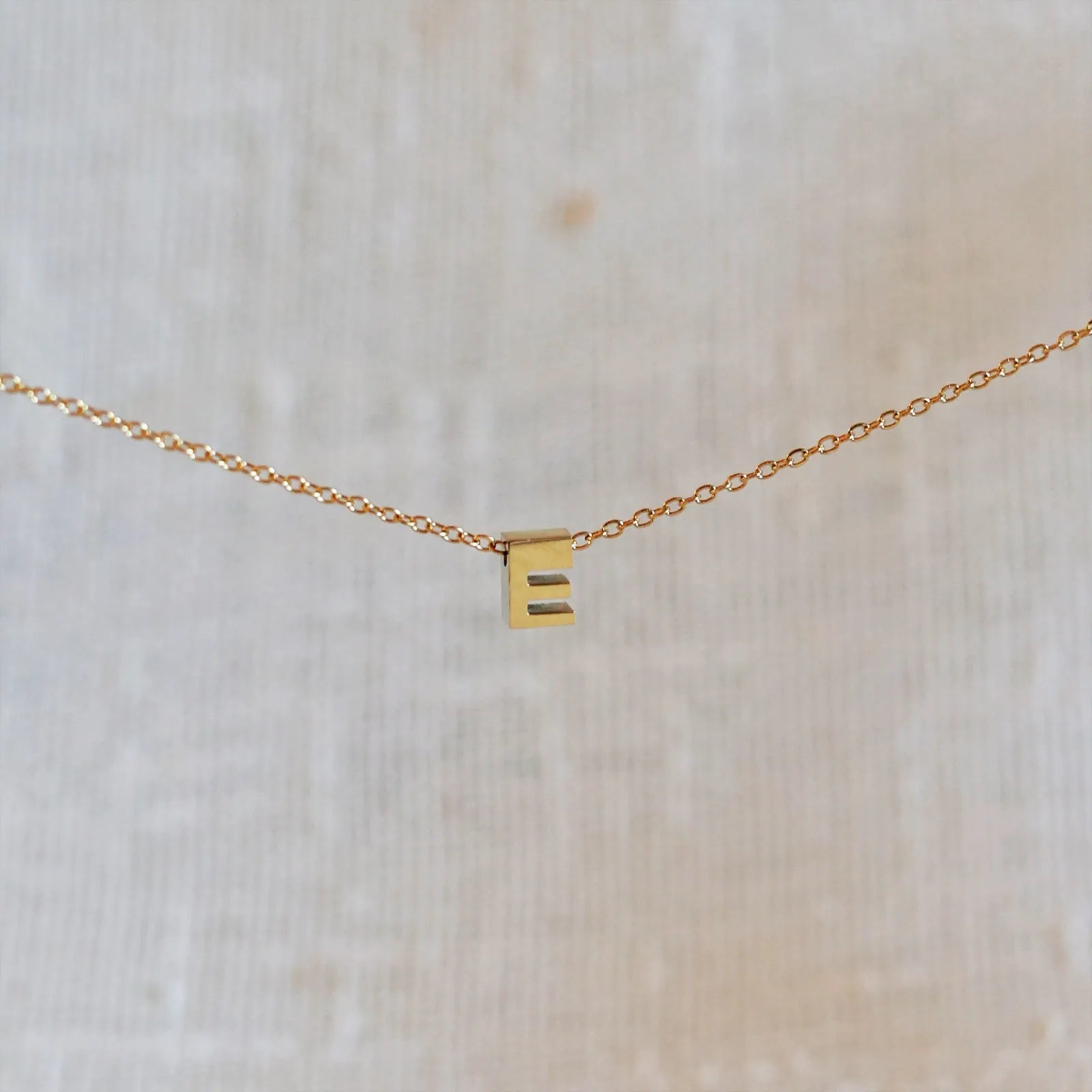 Collier L'instant avec pendentif lettre E doré en acier inoxydable, suspendu au centre d'une maille fine sur fond beige.