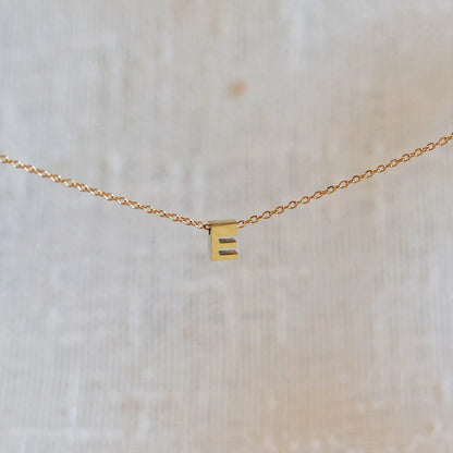 Collier L'instant avec pendentif lettre E doré en acier inoxydable, suspendu au centre d'une maille fine sur fond beige.