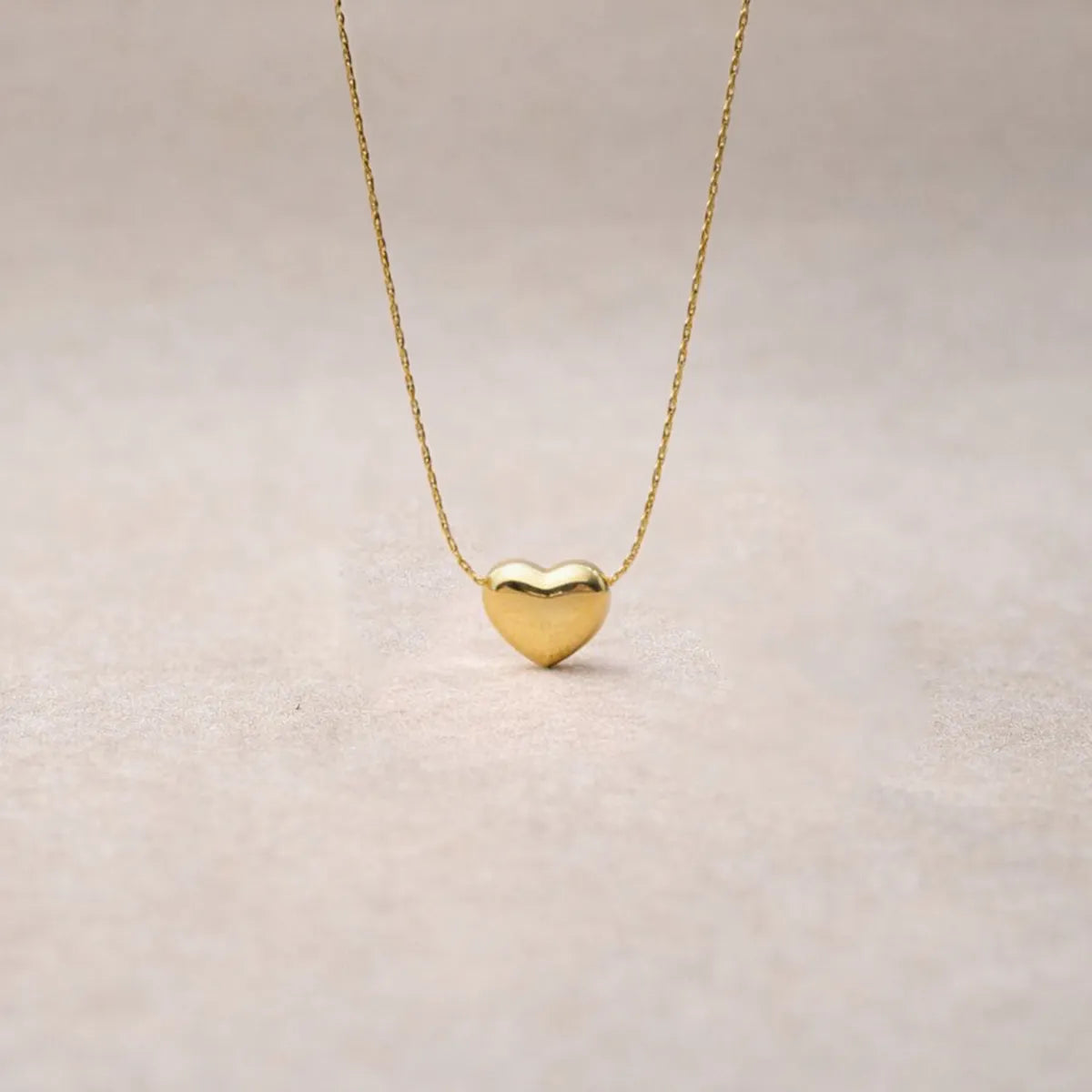 Collier Authentique avec pendentif cœur en acier inoxydable doré, photographié en gros plan sur un fond beige texturé.