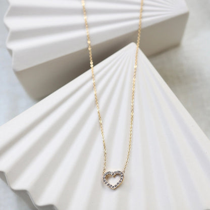 Collier Sentiment, pendentif cœur serti de brillants en acier inoxydable doré, posé sur un support beige texturé.