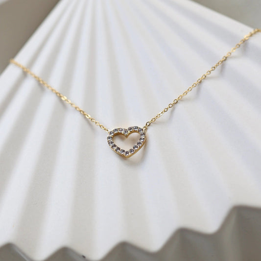 Collier Sentiment, pendentif cœur serti de brillants en acier inoxydable doré, photographié en gros plan sur un support beige texturé.