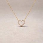 Collier Sentiment, pendentif cœur serti de brillants en acier inoxydable doré, photographié en gros plan sur un fond beige texturé.