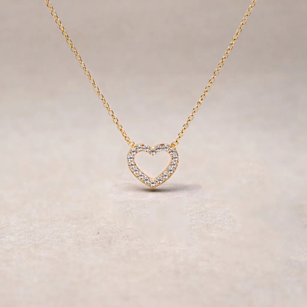 Collier Sentiment, pendentif cœur serti de brillants en acier inoxydable doré, photographié en gros plan sur un fond beige texturé.