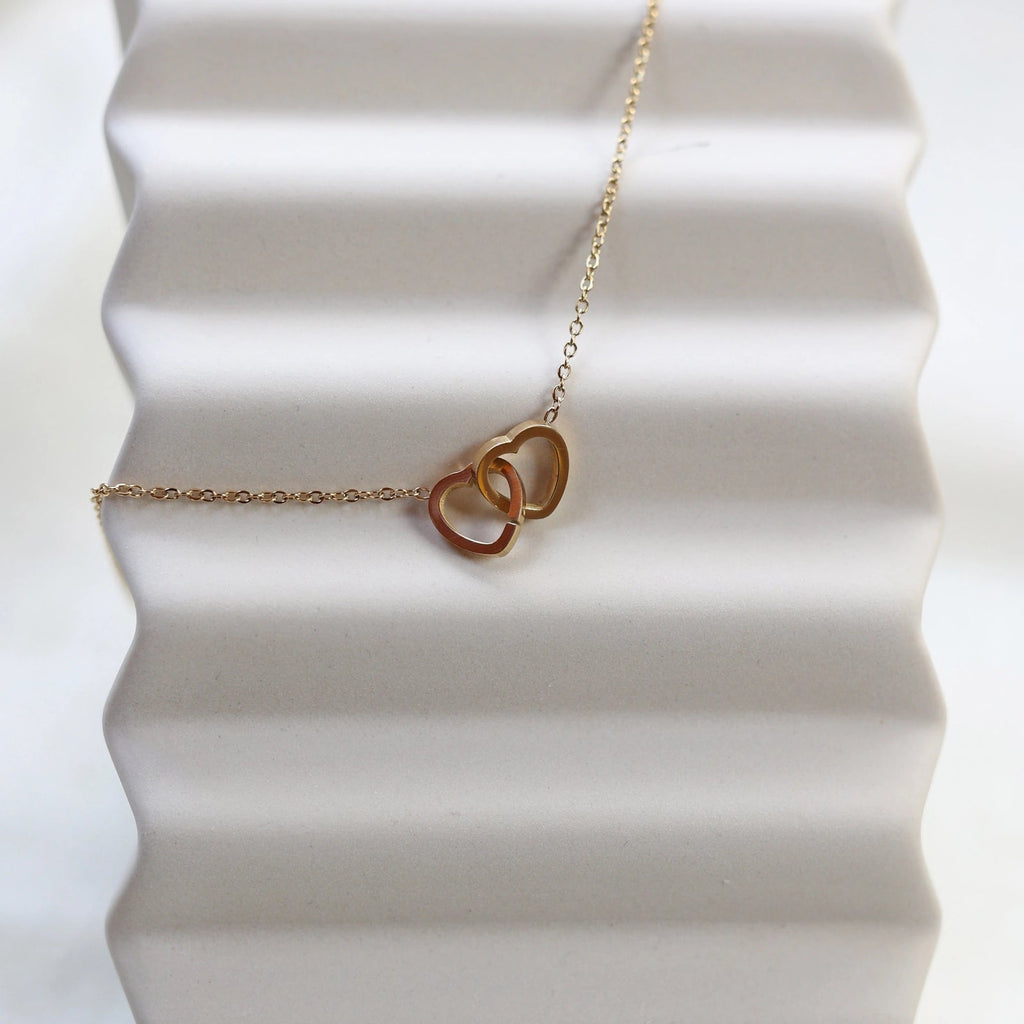 Collier Toi et moi en acier inoxydable doré, deux coeurs entrelacés, présenté en gros plan sur fond beige texturé.