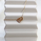 Collier Toi et moi en acier inoxydable doré, deux coeurs entrelacés, présenté en gros plan sur fond beige texturé.