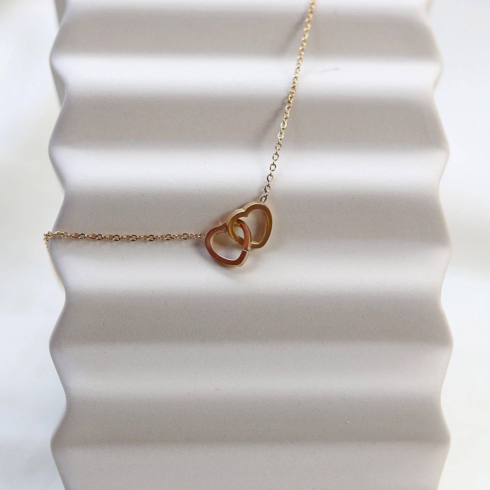 Collier Toi et moi en acier inoxydable doré, deux coeurs entrelacés, présenté en gros plan sur fond beige texturé.