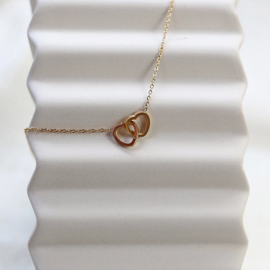Collier Toi et moi en acier inoxydable doré, deux coeurs entrelacés, présenté en gros plan sur fond beige texturé.