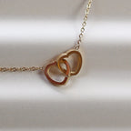 Collier Toi et moi en acier inoxydable doré, deux coeurs entrelacés, présenté en gros plan sur fond beige texturé.