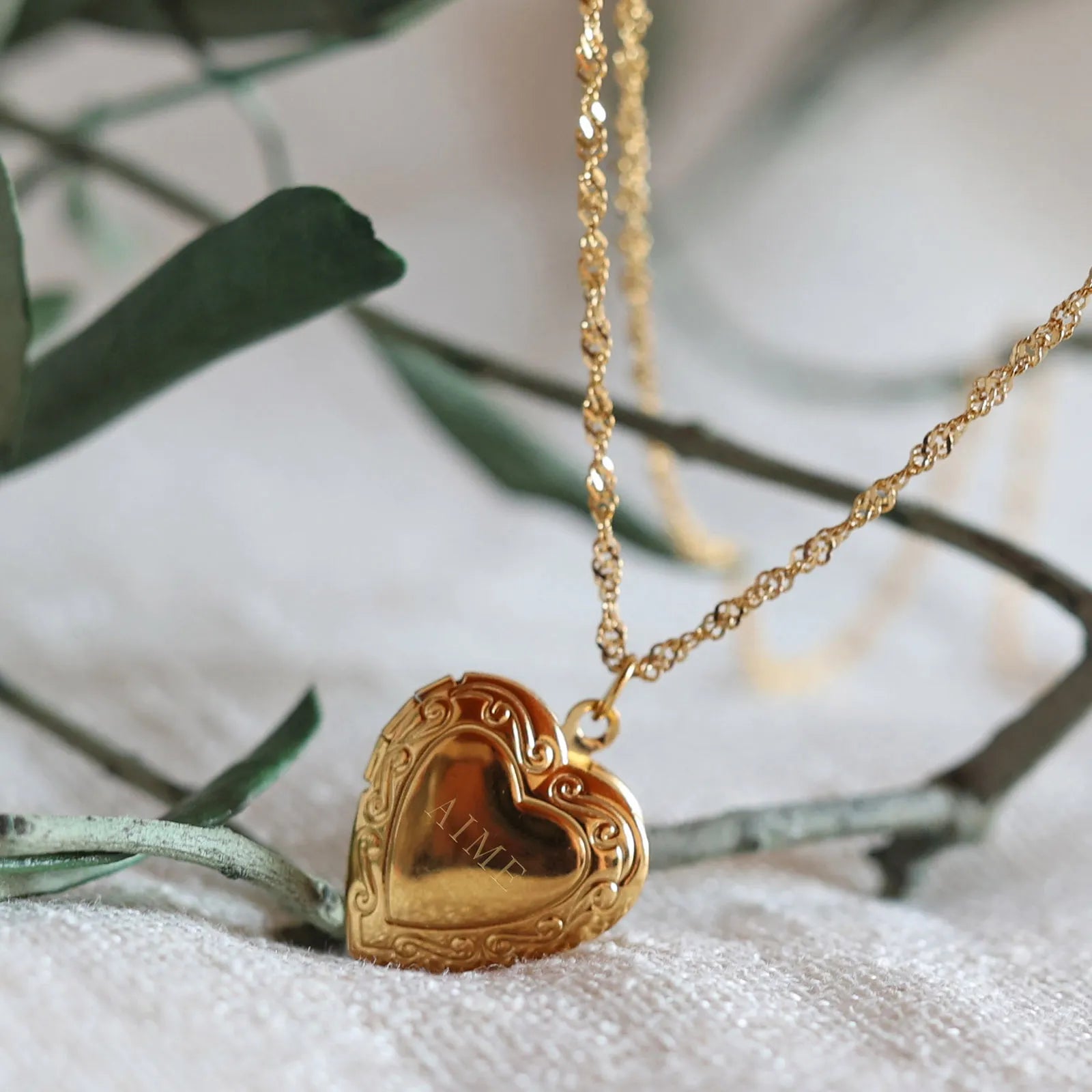 Zoom sur le collier "Entre nous", médaillon cœur en acier doré avec photo souvenir à personnaliser, gravé AIME posé sur un fond beige texturé.