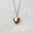 Zoom sur le collier "Entre nous", médaillon cœur en acier doré avec photo souvenir à personnaliser, posé sur un fond beige texturé.