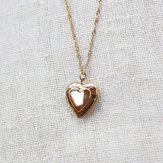 Zoom sur le collier "Entre nous", médaillon cœur en acier doré avec photo souvenir à personnaliser, posé sur un fond beige texturé.
