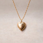 Zoom sur le collier "Entre nous", médaillon cœur en acier doré avec photo souvenir à personnaliser, posé sur un fond beige texturé.