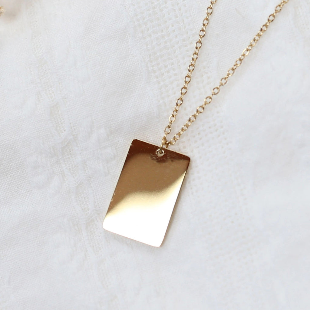 Zoom sur le collier "Je me souviens, en acier doré, avec médaille rectangulaire à graver, posé sur un fond beige.