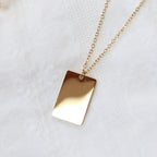 Zoom sur le collier "Je me souviens, en acier doré, avec médaille rectangulaire à graver, posé sur un fond beige.