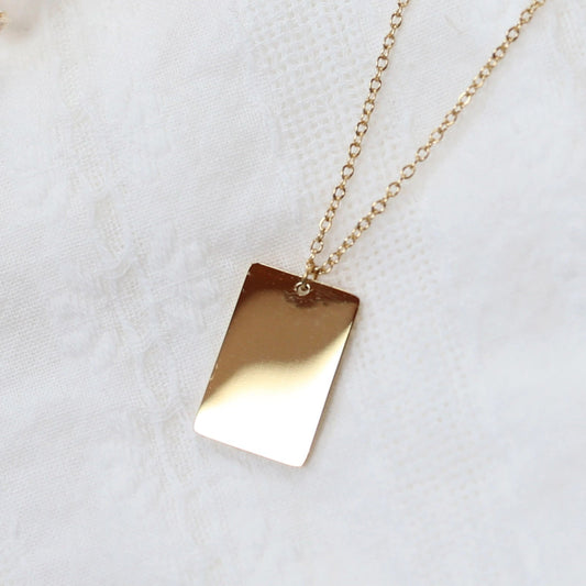Zoom sur le collier "Je me souviens, en acier doré, avec médaille rectangulaire à graver, posé sur un fond beige.