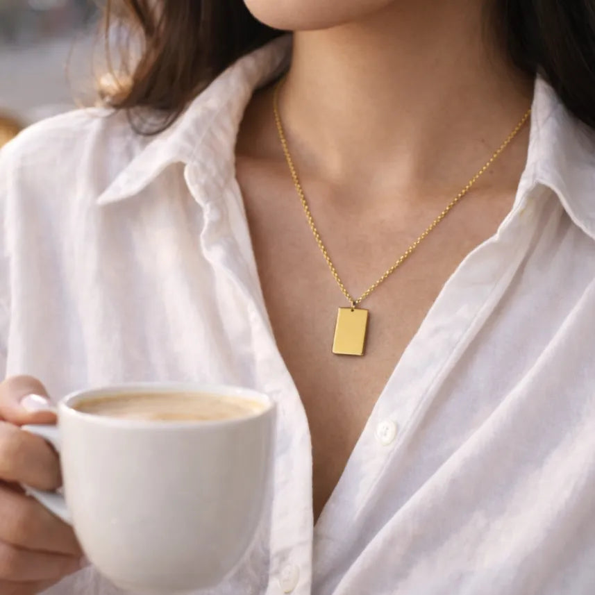 Collier Tracé en acier doré, avec médaille rectangulaire , porté autour du cou. Le modèle porte une chemise blanche et boit une tasse de café.