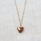 Collier "Entre nous", médaillon cœur en acier doré avec photo souvenir à personnaliser, posé sur un fond beige texturé.