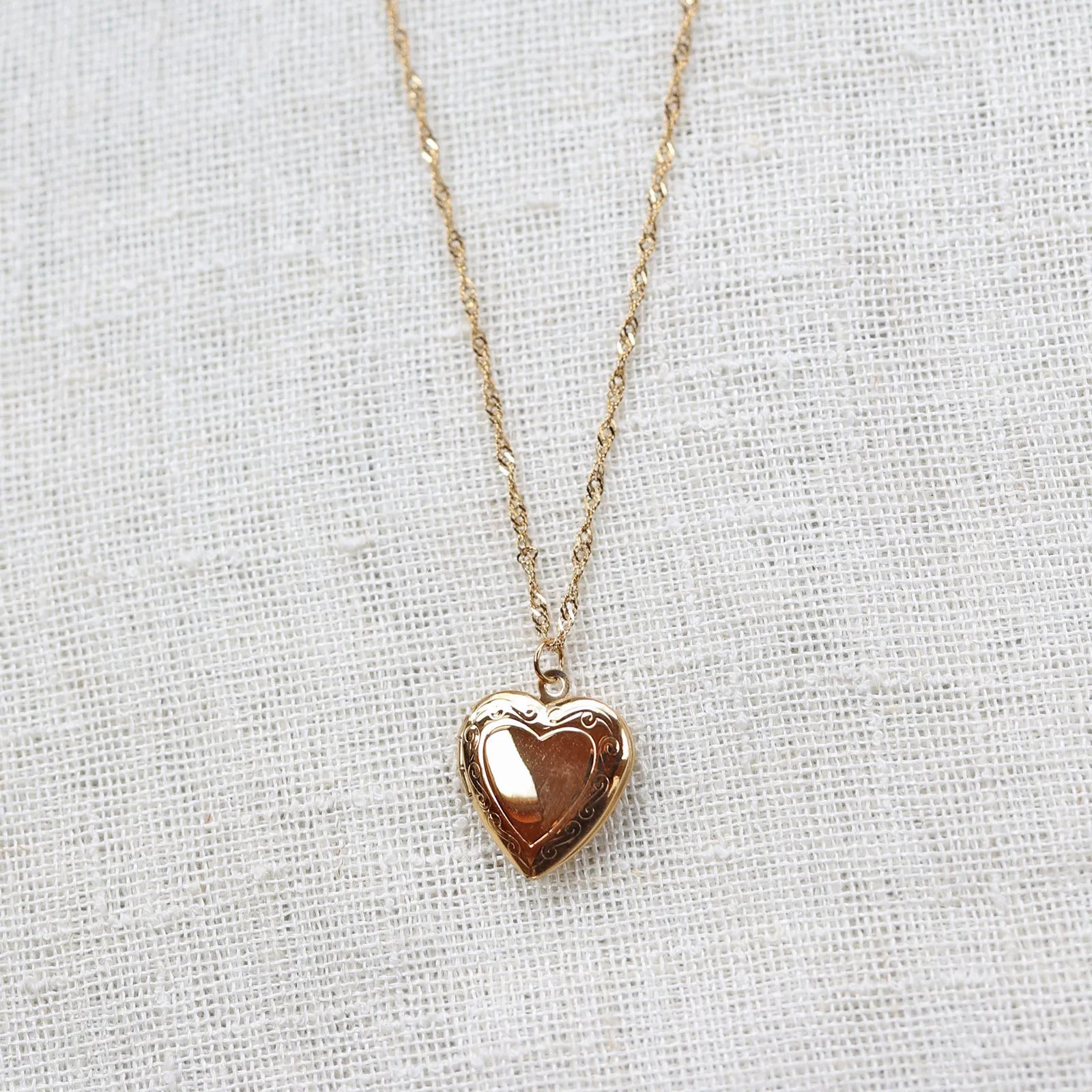 Collier "Entre nous", médaillon cœur en acier doré avec photo souvenir à personnaliser, posé sur un fond beige texturé.