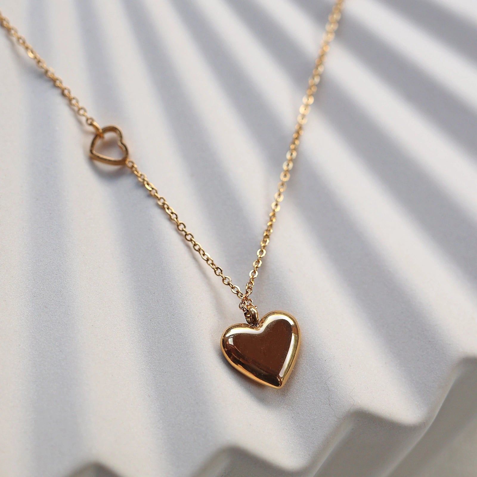 Zoom sur le collier "Ma vie" en acier doré, avec pendentif cœur personnalisable et petit coeur sur la chaîne, posé sur fond beige. 