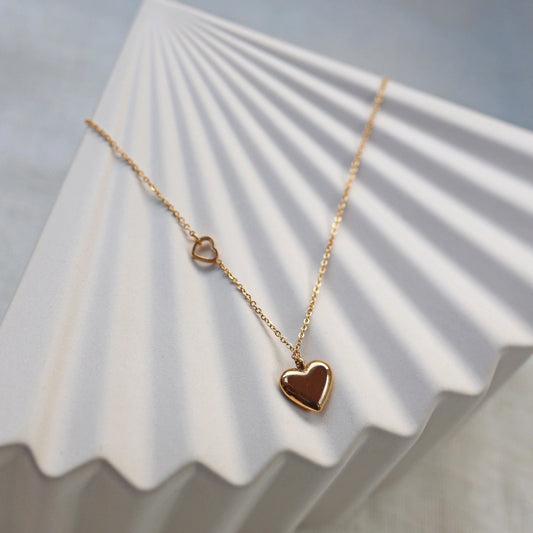 Collier "Ma vie" en acier doré, avec pendentif cœur personnalisable et petit coeur sur la chaîne, posé sur fond beige. 