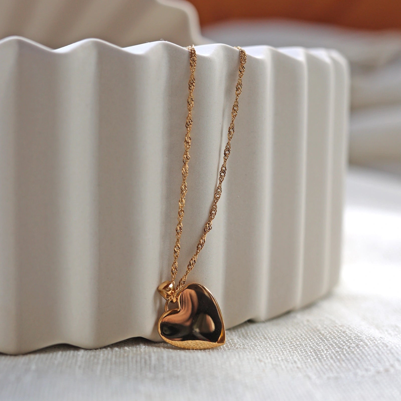 Collier "Pensée" en acier doré avec pendentif coeur strié orné d'un zircon central, personnalisable, posé de dos sur un fond beige texturé.
