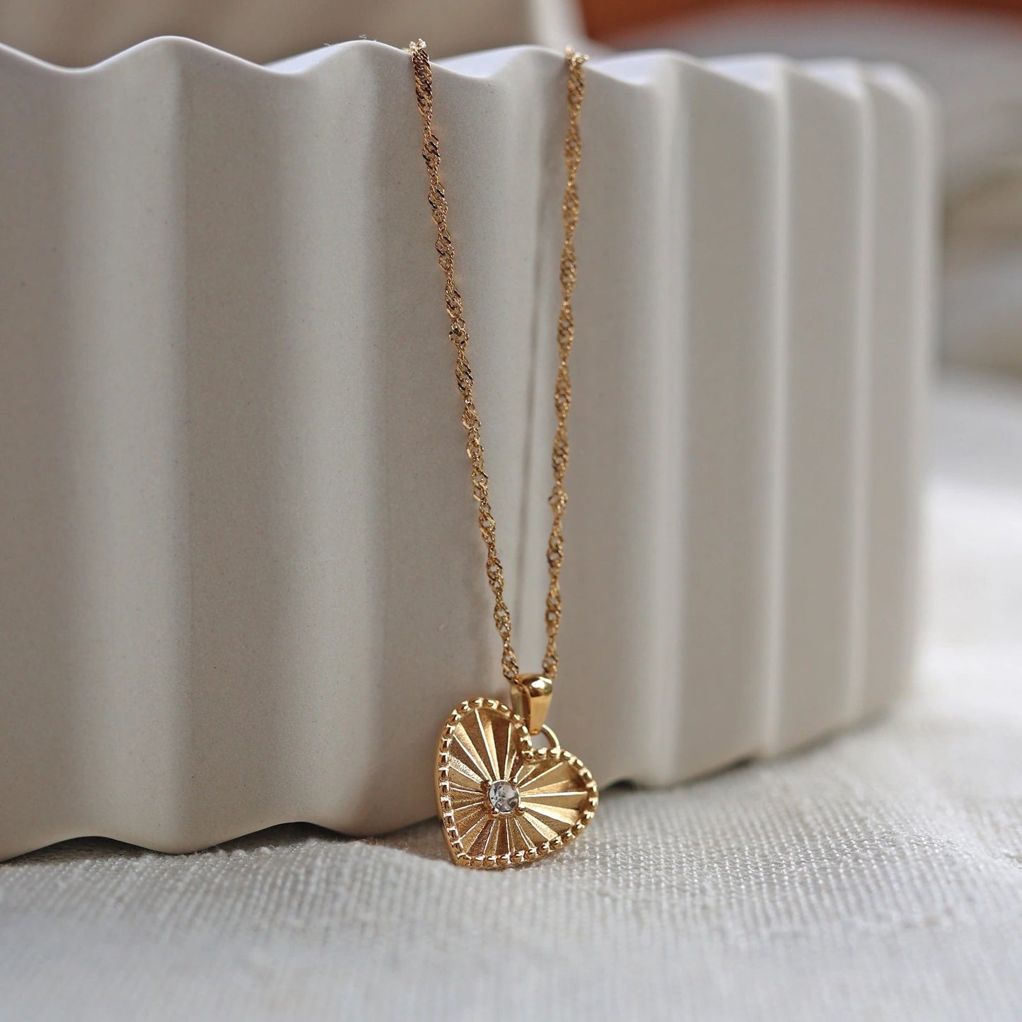 Collier "Pensée" en acier doré avec pendentif coeur strié orné d'un zircon central, personnalisable, posé sur un fond beige texturé.