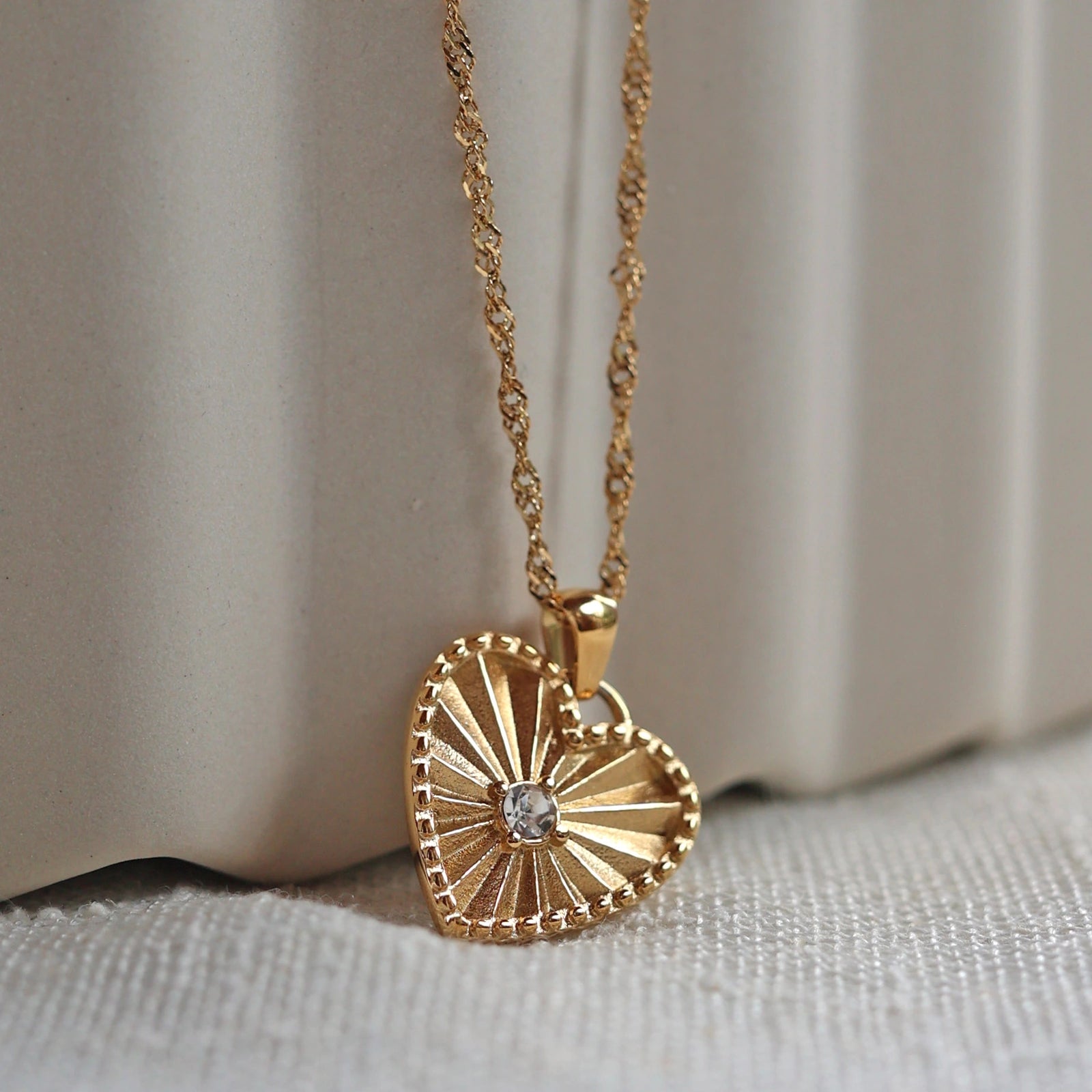Zoom sur le collier "Pensée" en acier doré avec pendentif coeur strié orné d'un zircon central, personnalisable, posé sur un fond beige texturé.