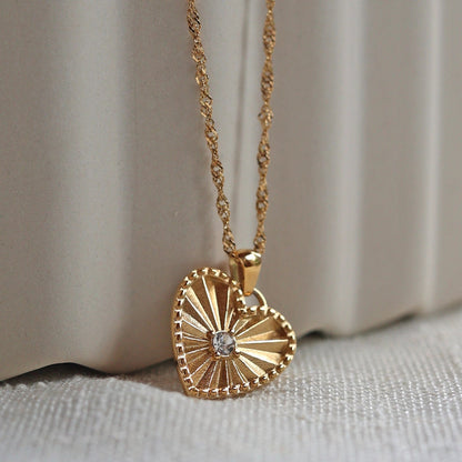 Zoom sur le collier "Pensée" en acier doré avec pendentif coeur strié orné d'un zircon central, personnalisable, posé sur un fond beige texturé.