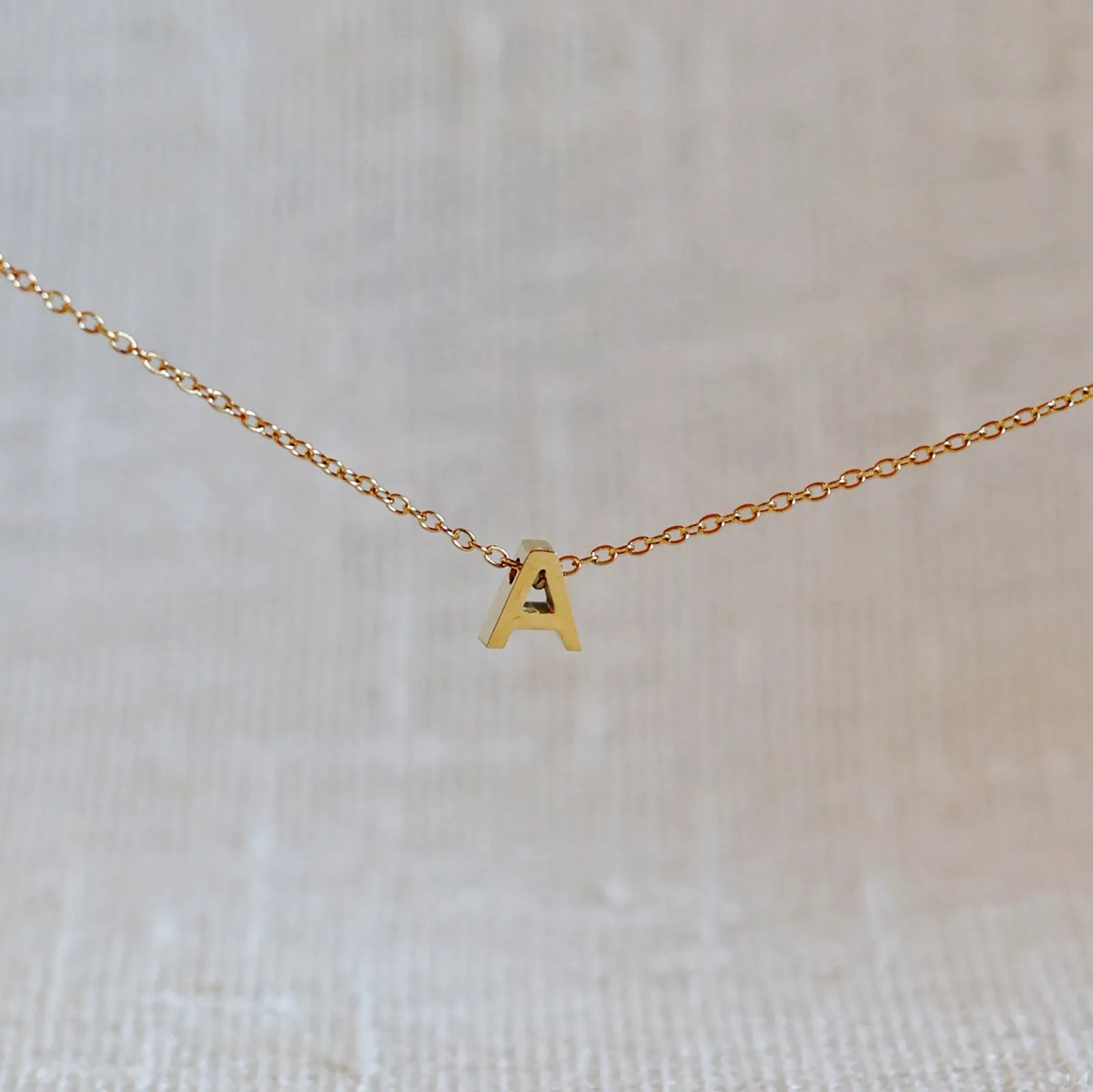 Collier L'instant avec pendentif lettre A en acier inoxydable doré, bijou minimaliste suspendu sur fond beige.