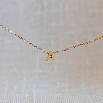 Collier L'instant avec pendentif lettre A en acier inoxydable doré, bijou minimaliste suspendu sur fond beige.