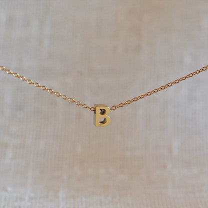 Collier L'instant avec pendentif lettre B doré en acier inoxydable, suspendu au centre d'une maille fine sur fond beige.