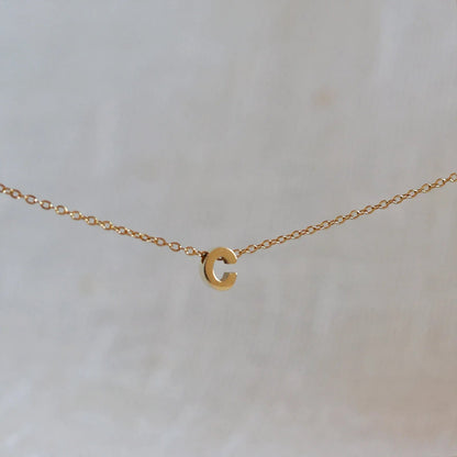Collier L'instant avec pendentif lettre C doré en acier inoxydable, suspendu au centre d'une maille fine sur fond beige.