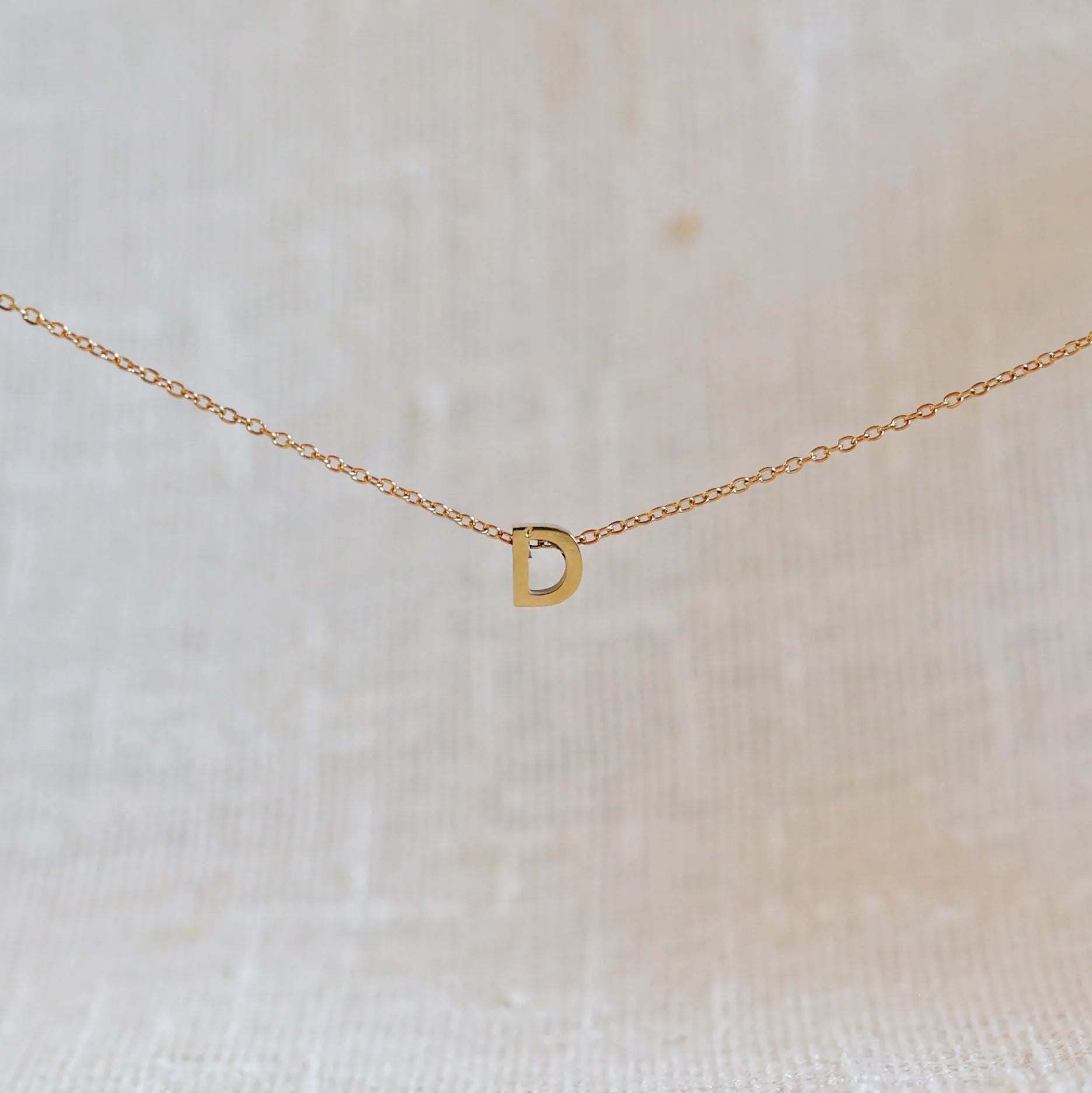 Collier L'instant avec pendentif lettre D doré en acier inoxydable, suspendu au centre d'une maille fine sur fond beige.