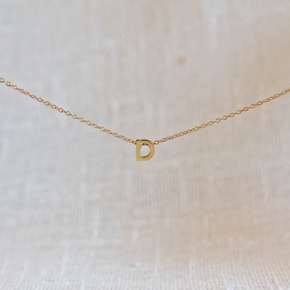 Collier L'instant avec pendentif lettre D doré en acier inoxydable, suspendu au centre d'une maille fine sur fond beige.
