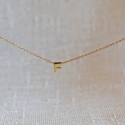 Collier L'instant avec pendentif lettre F doré en acier inoxydable, suspendu au centre d'une maille fine sur fond beige.