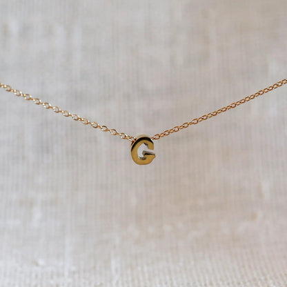 Collier L'instant avec pendentif lettre G doré en acier inoxydable, suspendu au centre d'une maille fine sur fond beige.