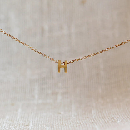 Collier L'instant avec pendentif lettre H doré en acier inoxydable, suspendu au centre d'une maille fine sur fond beige.