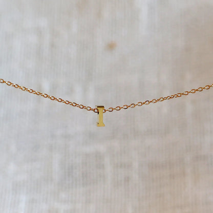 Collier L'instant avec pendentif lettre I doré en acier inoxydable, suspendu au centre d'une maille fine sur fond beige.