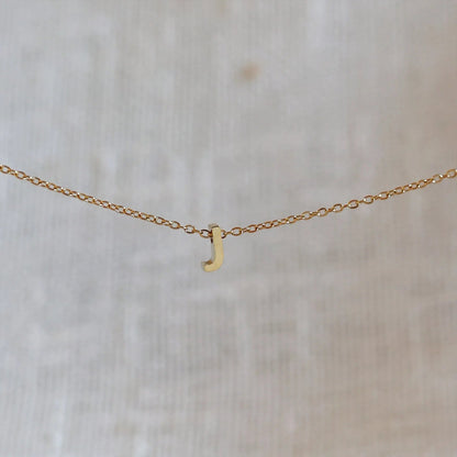 Collier L'instant avec pendentif lettre J doré en acier inoxydable, suspendu au centre d'une maille fine sur fond beige.